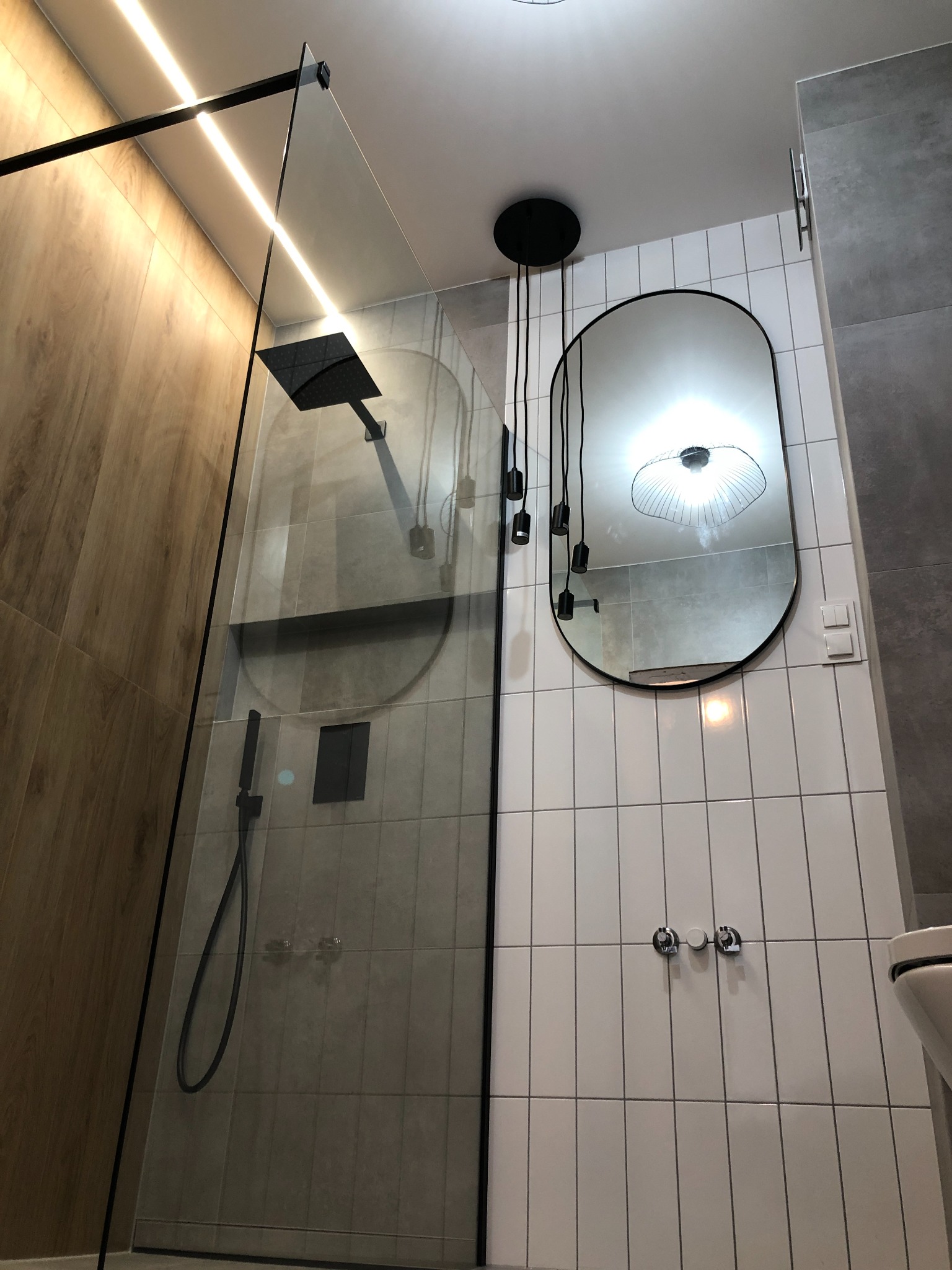 Nowoczesna łazienka z kabiną prysznicową typu walk-in z czarnymi profilami, owalnym lustrem z czarną ramą i wiszącą lampą z kilkoma czarnymi kloszami na przewodach.