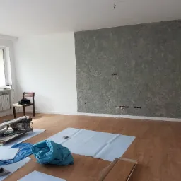 Pokój po remoncie z dekoracyjną szarą ścianą, widoczne przygotowane narzędzia i materiały do dalszych prac wykończeniowych, podłoga zabezpieczona folią ochronną.