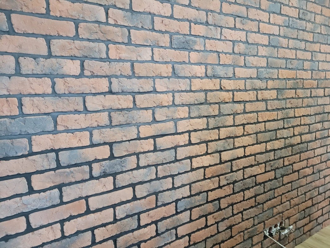 Ściana z dekoracyjnej cegły w odcieniach pomarańczy i szarości, z widocznym gniazdkiem elektrycznym w dolnym rogu.