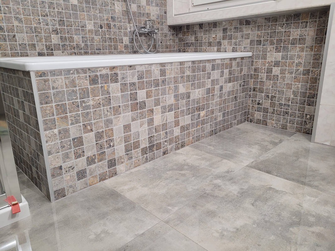 Wykończona łazienka z mozaiką w odcieniach szarości i brązu na obudowie wanny i ścianach, oraz szare płytki podłogowe.