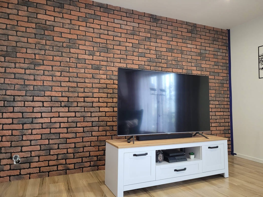 Ściana z cegły klinkierowej w salonie, z widocznym gniazdkiem elektrycznym po lewej stronie i telewizorem na białej szafce RTV.
