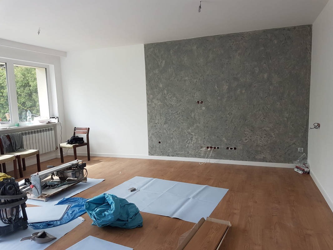 Pokój po remoncie z dekoracyjną szarą ścianą, widoczne przygotowane narzędzia i materiały do dalszych prac wykończeniowych, podłoga zabezpieczona folią ochronną.