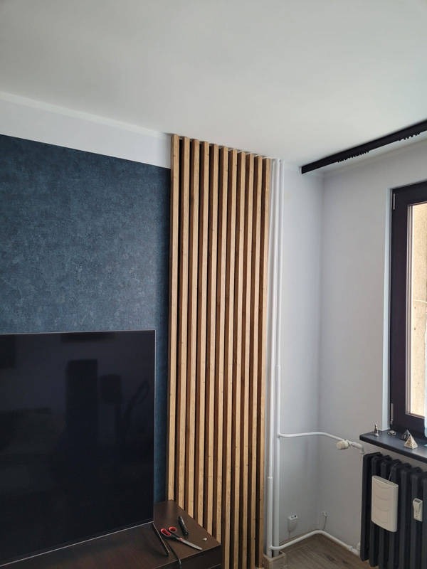 Drewniane lamele dekoracyjne na ścianie w salonie, obok telewizora i grzejnika, widoczne rury instalacyjne.