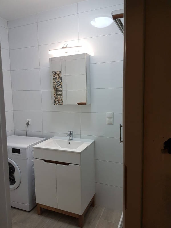 Biała łazienka z pralką, umywalką z szafką i lustrem z oświetleniem, wykończona białymi płytkami. Fragment dekoracyjnej płytki na lustrze. Widok z otwartych drzwi.