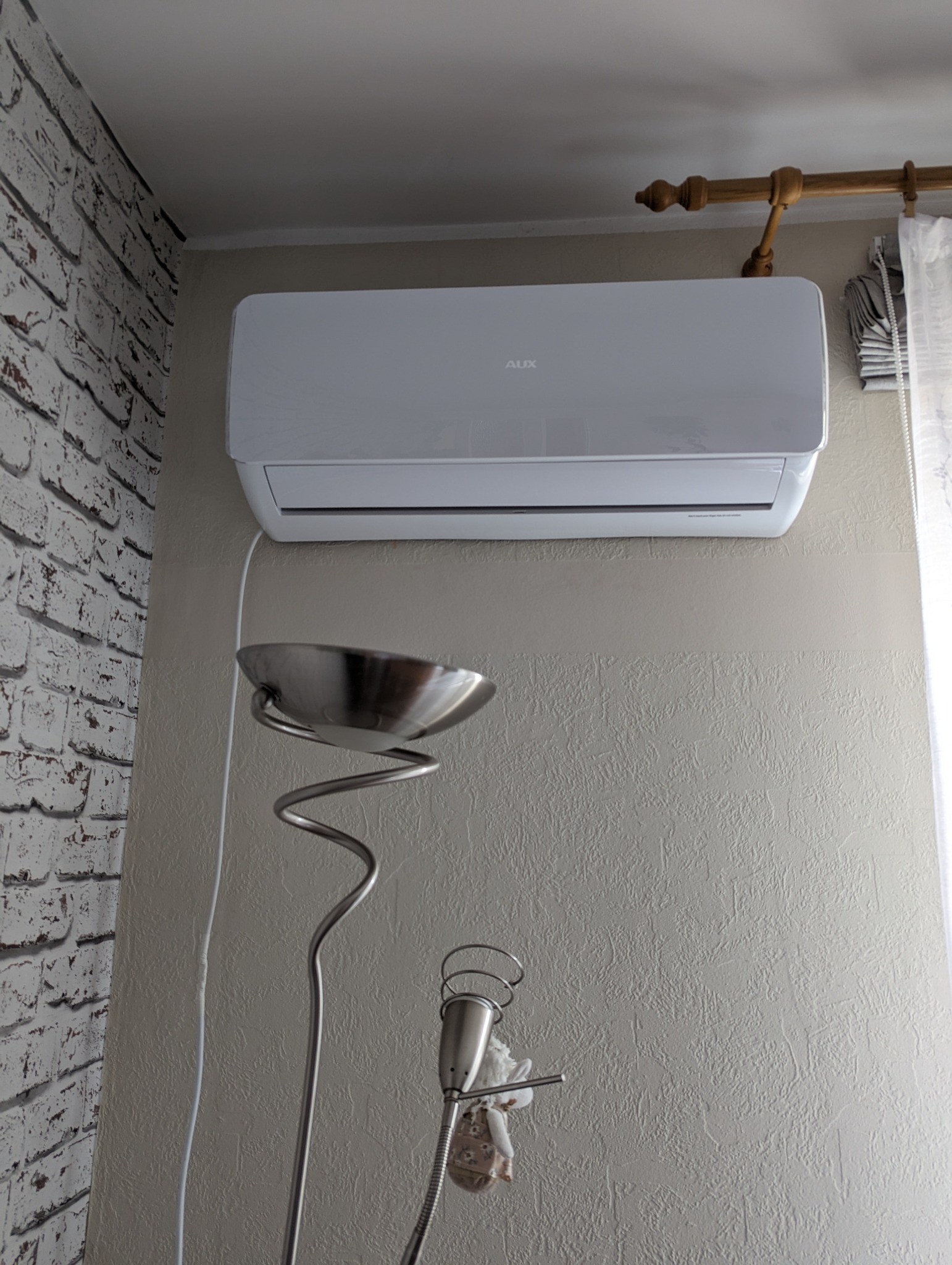 Biały klimatyzator marki AUX zamontowany na ścianie w pokoju, widoczna lampa podłogowa i fragment ściany z cegły.