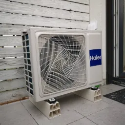 Zewnętrzna jednostka klimatyzacji Haier zamontowana na specjalnych podstawach na zewnątrz budynku, widoczna drewniana elewacja i okno.