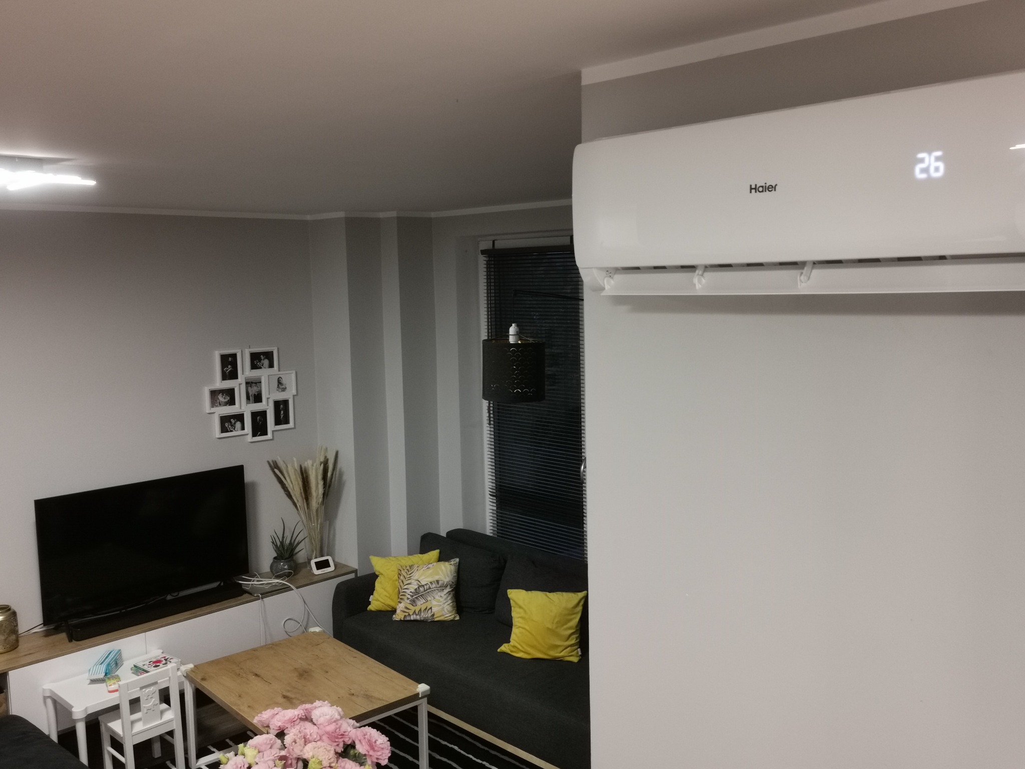 Biały klimatyzator Haier zamontowany na ścianie w szarym salonie, widoczny wyświetlacz z temperaturą 26 stopni Celsjusza, w tle sofa z żółtymi poduszkami i telewizor.