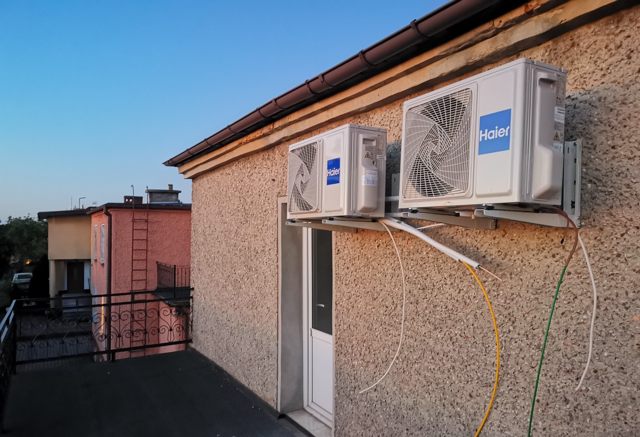 Dwa klimatyzatory Haier zamontowane na elewacji budynku z widocznymi przewodami instalacyjnymi, w tle inne budynki i błękitne niebo.