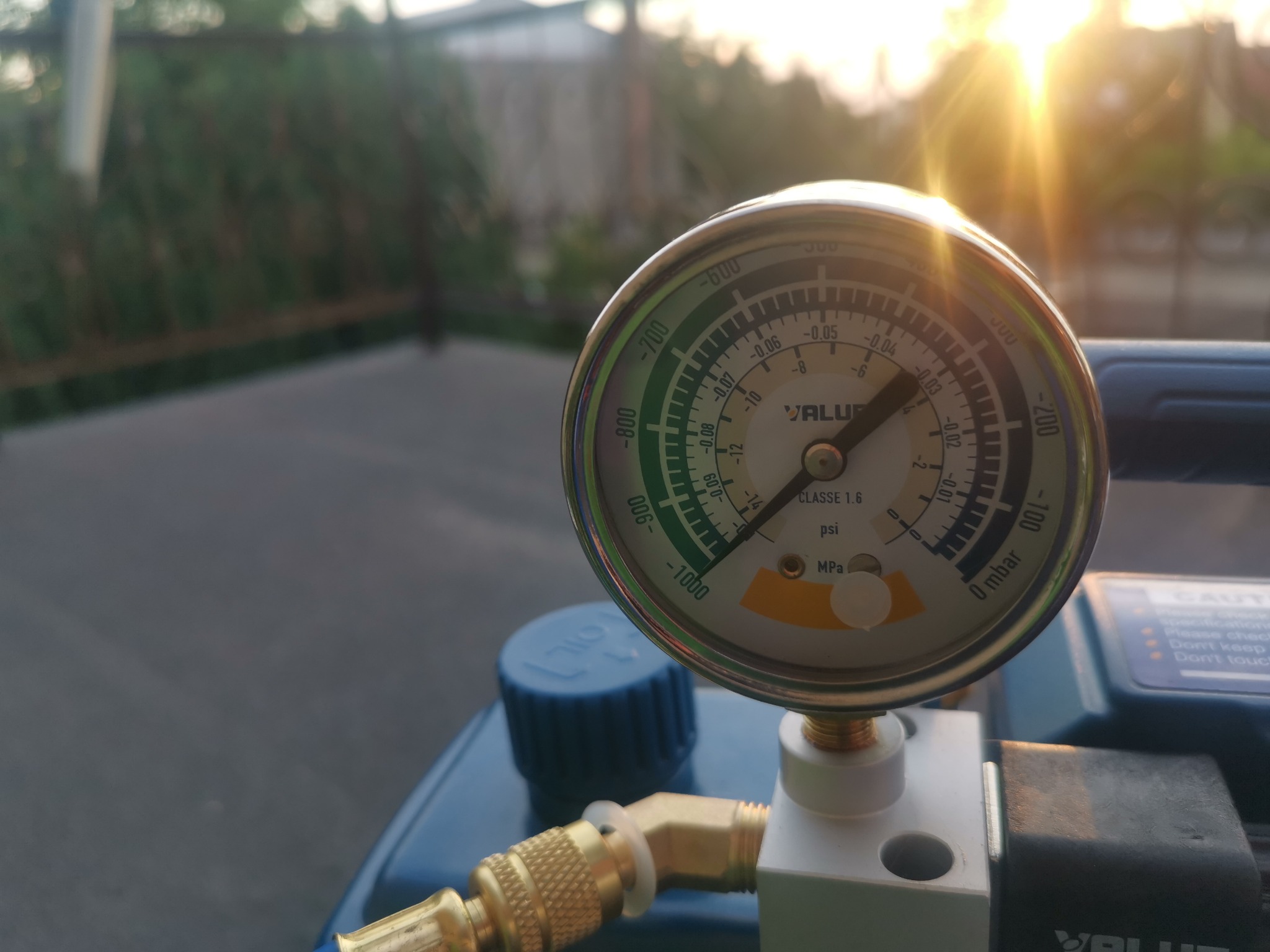 Manometr na niebieskiej pompie próżniowej VALUE, wskazujący podciśnienie, z widocznymi przyłączami mosiężnymi i słońcem w tle.