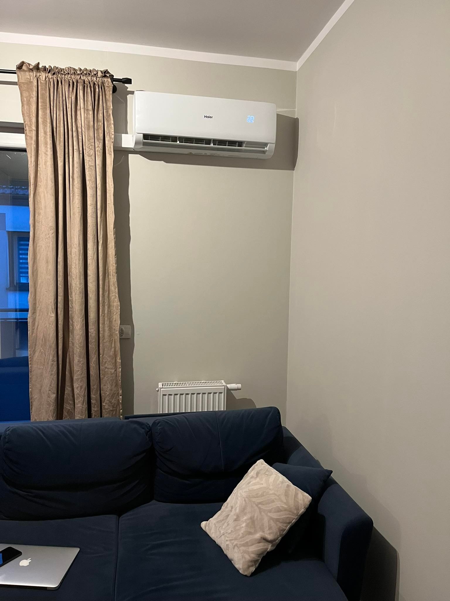 Biały klimatyzator Haier na szarej ścianie w pokoju dziennym nad niebieską sofą, obok zasłoniętego okna.