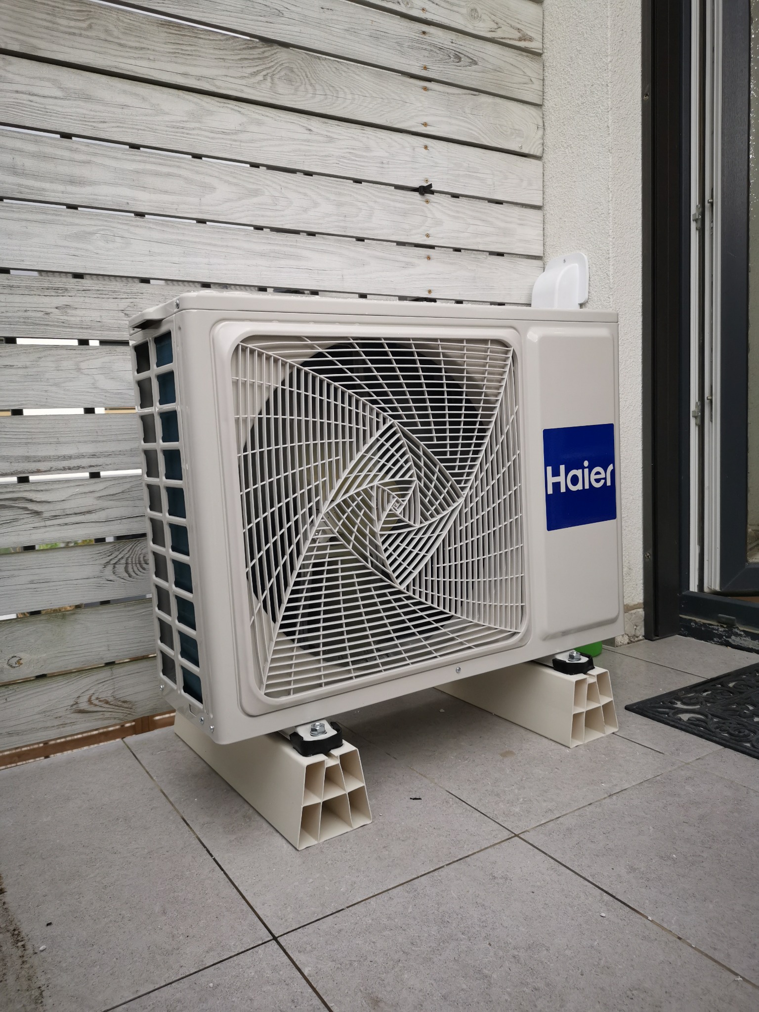 Zewnętrzna jednostka klimatyzacji Haier zamontowana na specjalnych podstawach na zewnątrz budynku, widoczna drewniana elewacja i okno.