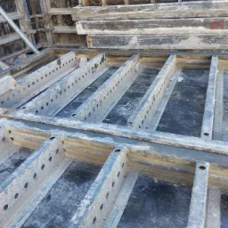 Szare, metalowe szalunki fundamentowe z widocznymi otworami, przygotowane do wylania betonu, z elementami rusztowania w tle.