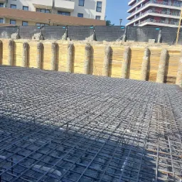 Siatka zbrojeniowa na czarnej folii, przygotowana do zalania betonem, z widocznym zabezpieczeniem wykopu deskami i żelbetowymi kolumnami, w tle budynki mieszkalne.