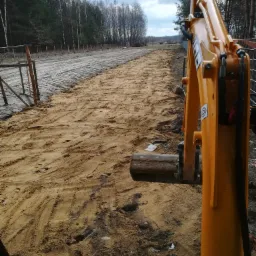 Żółta koparka na placu budowy, świeżo wyrównany teren z piaskiem, widoczne ślady opon i gąsienic, w tle las i pochmurne niebo.
