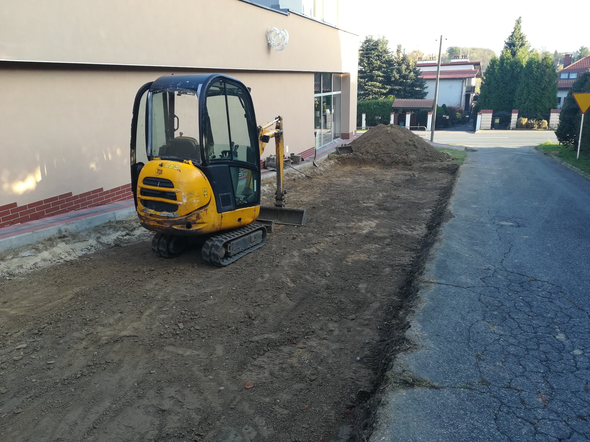 Żółta mini koparka JCB na placu budowy przed budynkiem, z wykopem wzdłuż drogi i stertą ziemi w tle.