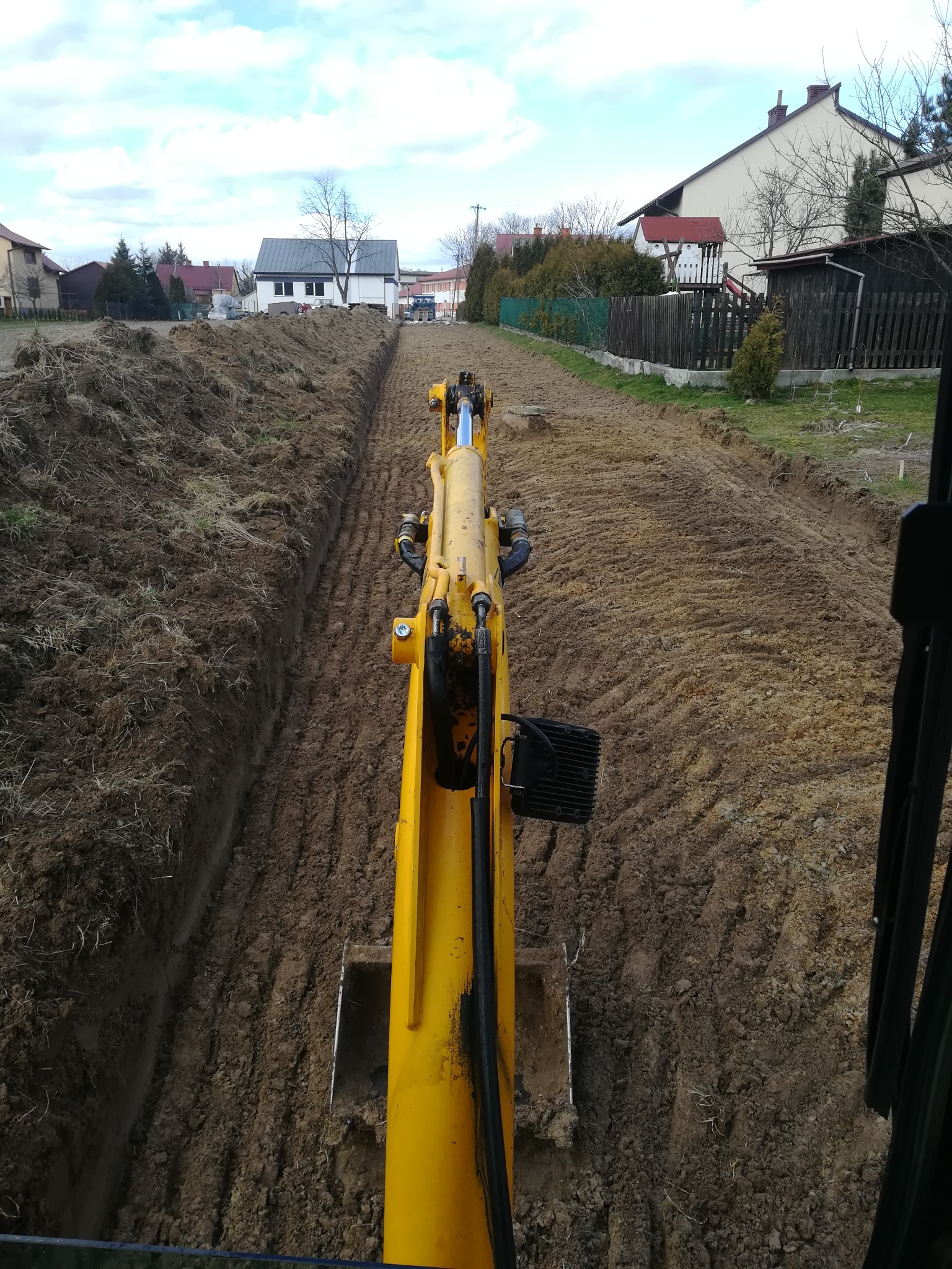 Żółta koparka widziana z perspektywy operatora, tworząca długi, wąski wykop w ziemi na tle zabudowań jednorodzinnych i pochmurnego nieba.