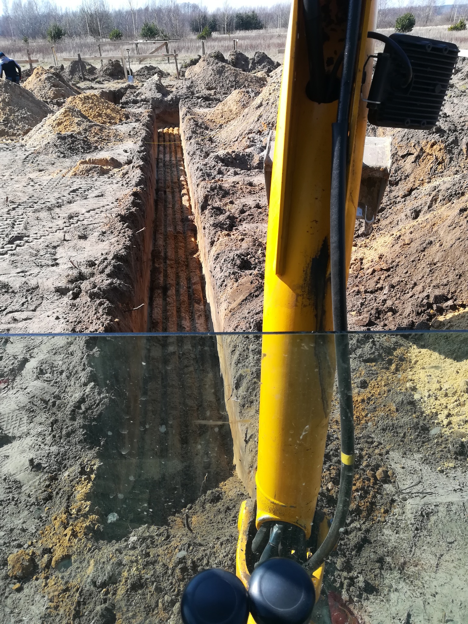 Żółta ramię koparki nad wykopem z ułożonymi rurami, widoczne ślady opon i stosy ziemi w tle, praca w toku.