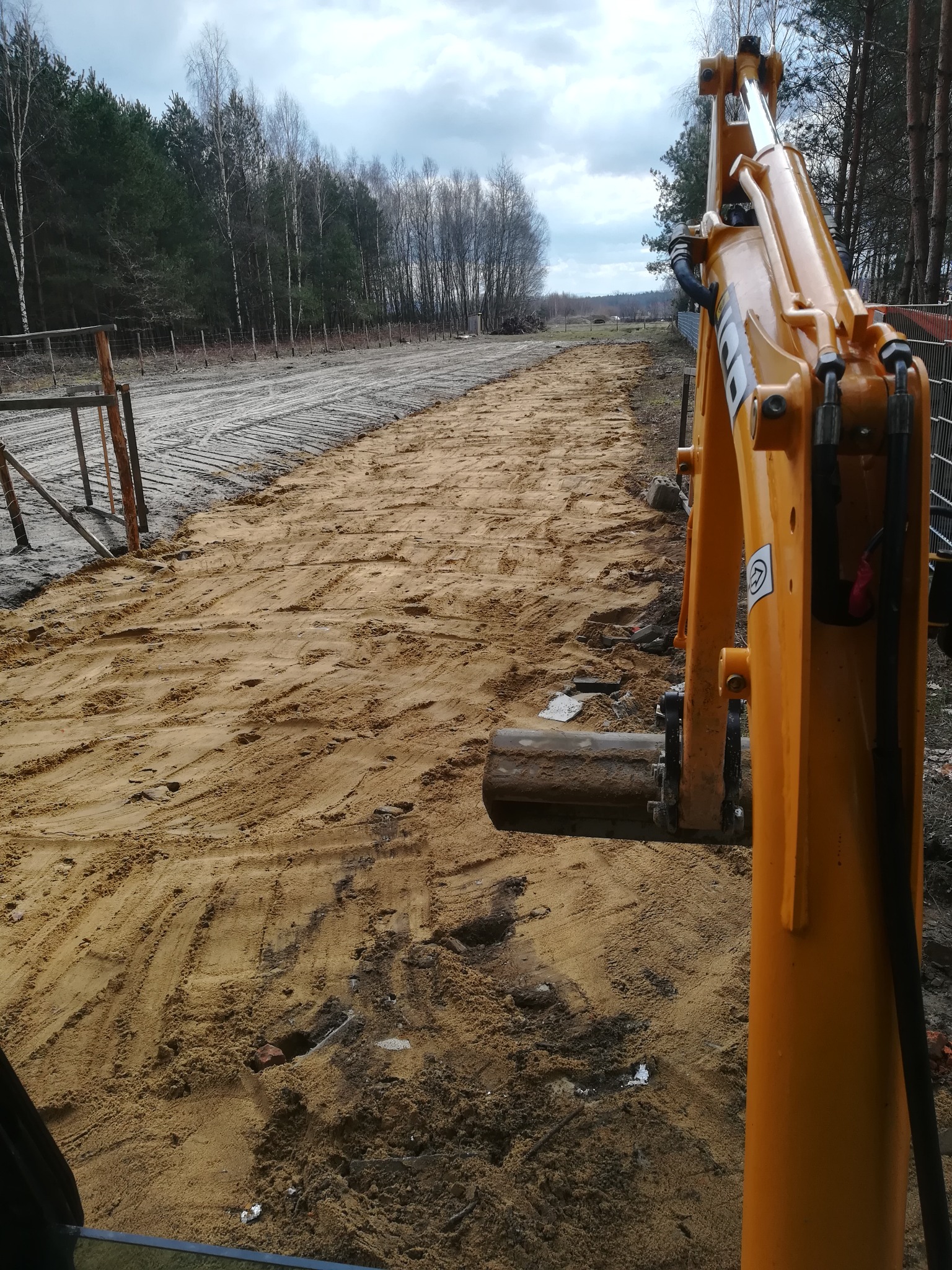 Żółta koparka na placu budowy, świeżo wyrównany teren z piaskiem, widoczne ślady opon i gąsienic, w tle las i pochmurne niebo.