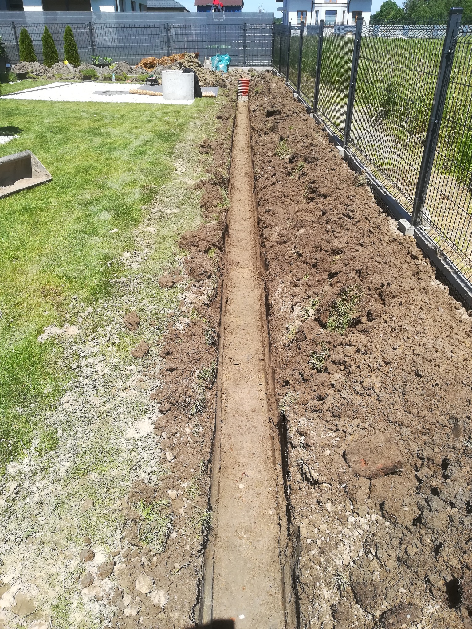 Wąski wykop w ziemi, widok z góry, w tle trawnik, ogrodzenie panelowe i dom jednorodzinny.