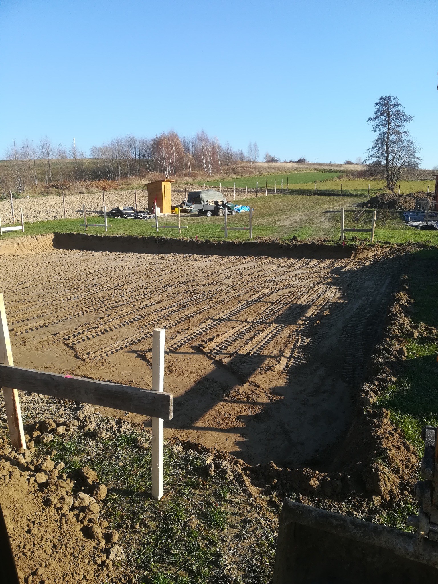 Przygotowany teren pod budowę z widocznymi śladami opon, drewniane słupki wyznaczające granice, w tle budka i sprzęt budowlany na tle zielonego pola i drzew.