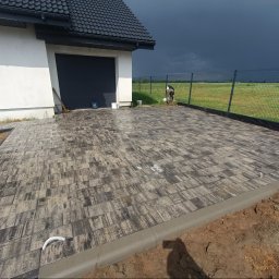 Art Bruk Joachim Jankowski - Nowa, szara kostka brukowa ułożona na podjeździe przed domem z garażem, obok metalowe ogrodzenie. Widoczne ciemne chmury na niebie.