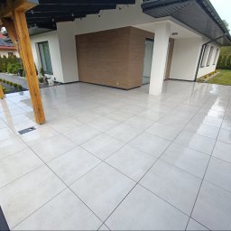 Art Bruk Joachim Jankowski - Nowoczesny taras z jasnych, kwadratowych płytek, drewniana pergola, czarne obrzeże, widok na dom i ogród. Dwóch mężczyzn w tle.