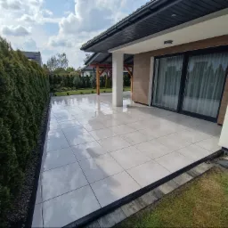 Jasnoszara, błyszcząca, kwadratowa podłoga z płytek na zewnątrz domu. Widoczny fragment ogrodu i żywopłot. Nowoczesny design i dbałość o detale.