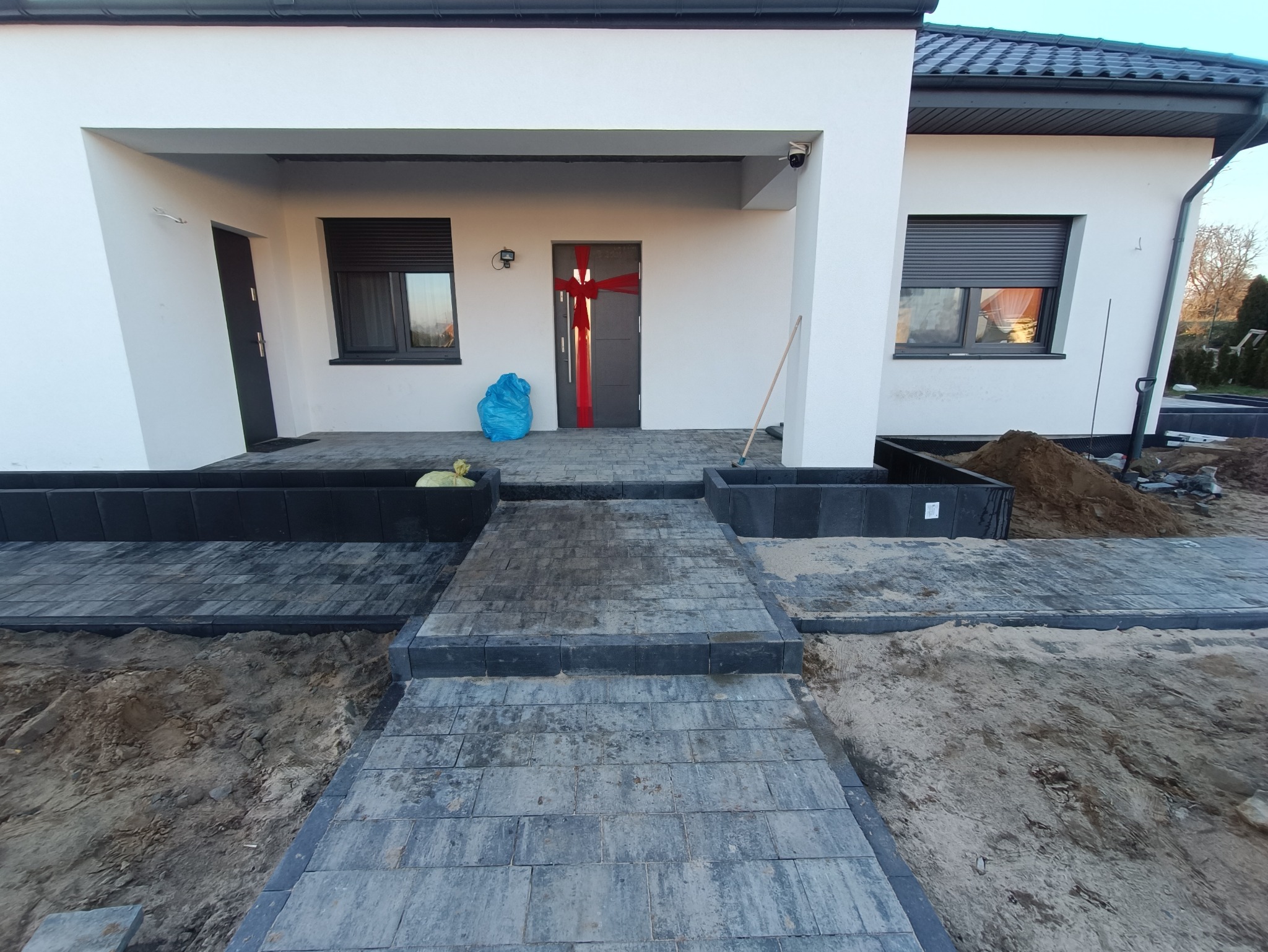 Ułożona kostka brukowa prowadząca do wejścia domu, ozdobionego czerwoną wstęgą. Widoczne obrzeża rabat i przygotowane podłoże.