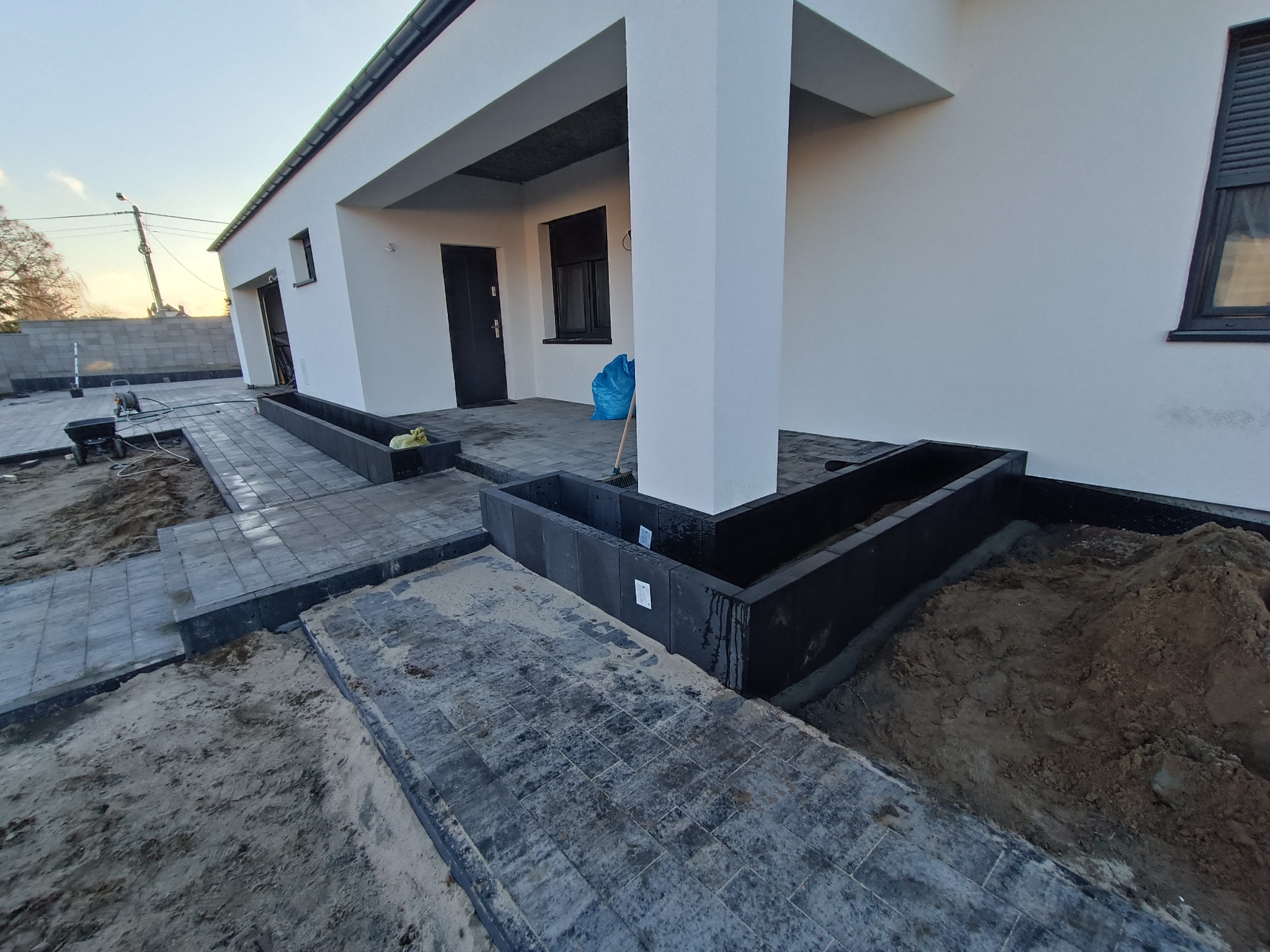 Ułożona kostka brukowa wzdłuż ścieżki i podjazdu domu z przygotowanymi rabatami kwiatowymi, widoczne ślady piasku i ziemi po pracach budowlanych.