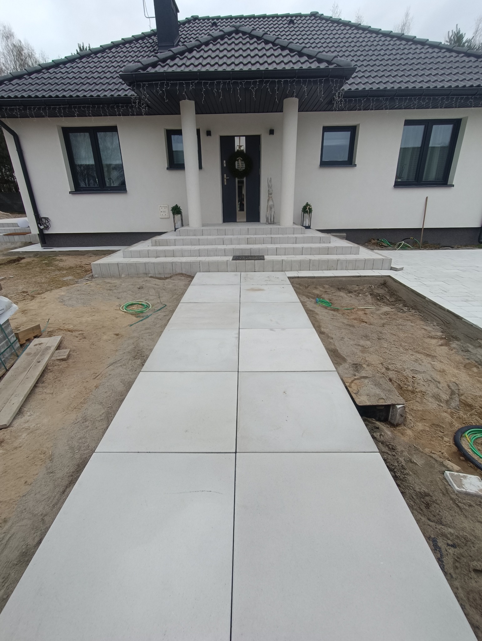 Ścieżka z dużych, szarych płyt prowadząca do wejścia do nowoczesnego domu z białymi ścianami i ciemną dachówką, widoczne przygotowania do dalszych prac w ziemi po bokach ścieżki.