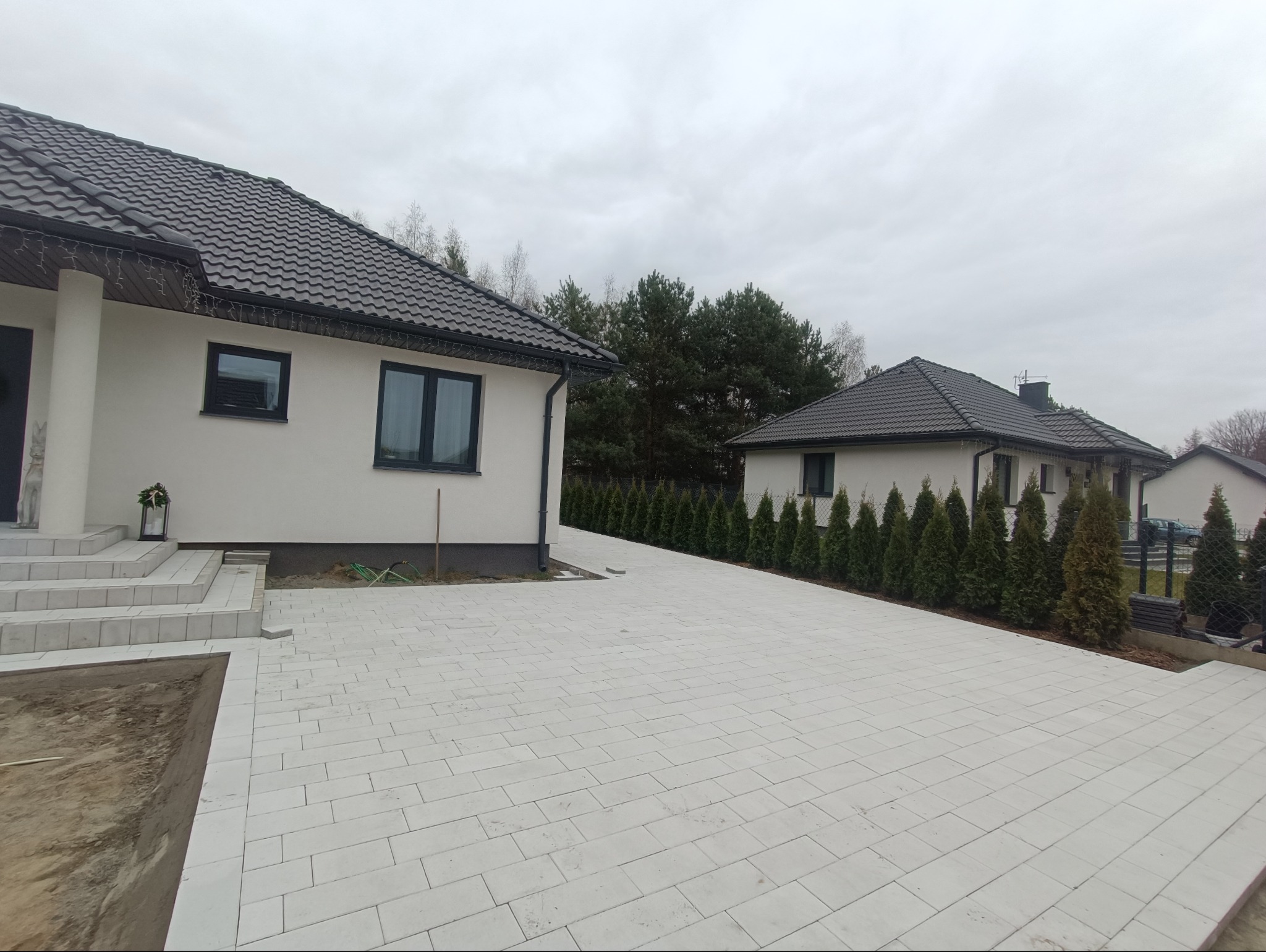 Nowoczesny podjazd z jasnoszarej kostki brukowej prowadzący do dwóch budynków mieszkalnych z ciemnymi dachami, obrzeża obsadzone tujami.