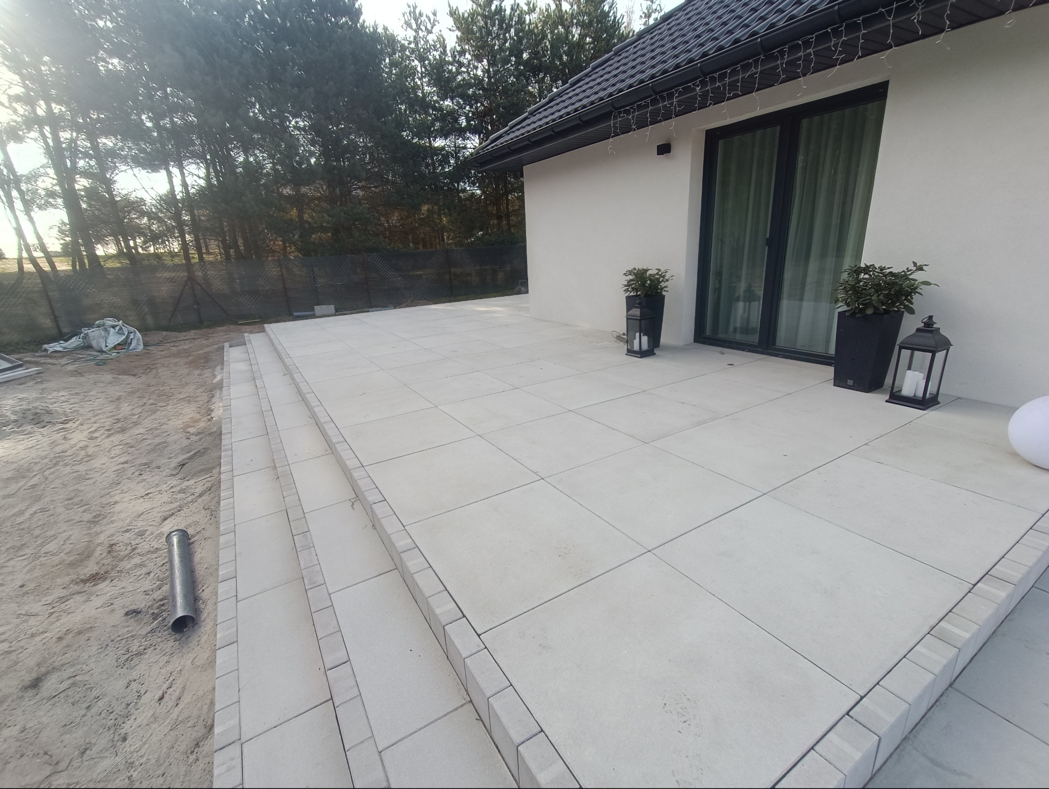 Jasna, nowa kostka brukowa na tarasie przed domem z czarnymi ramami okiennymi, dekoracyjnymi lampionami i roślinami w donicach, widoczne schody z obrzeżami i teren przygotowany pod dalsze prace.