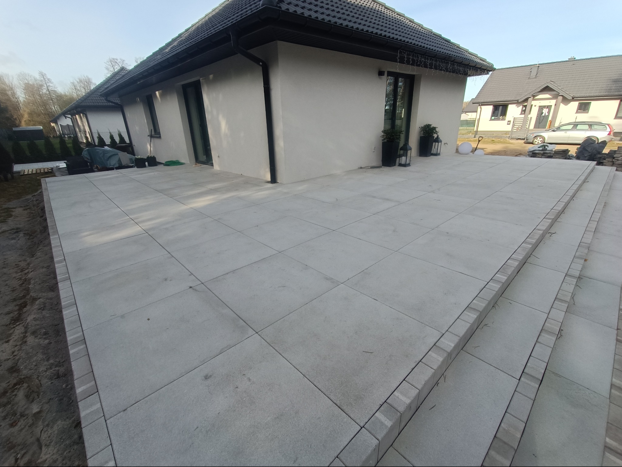 Rozległy, szary taras z płyt betonowych, prowadzący do nowoczesnego domu z ciemną dachówką, widok z niskiej perspektywy, z widocznymi schodami obłożonymi kostką.