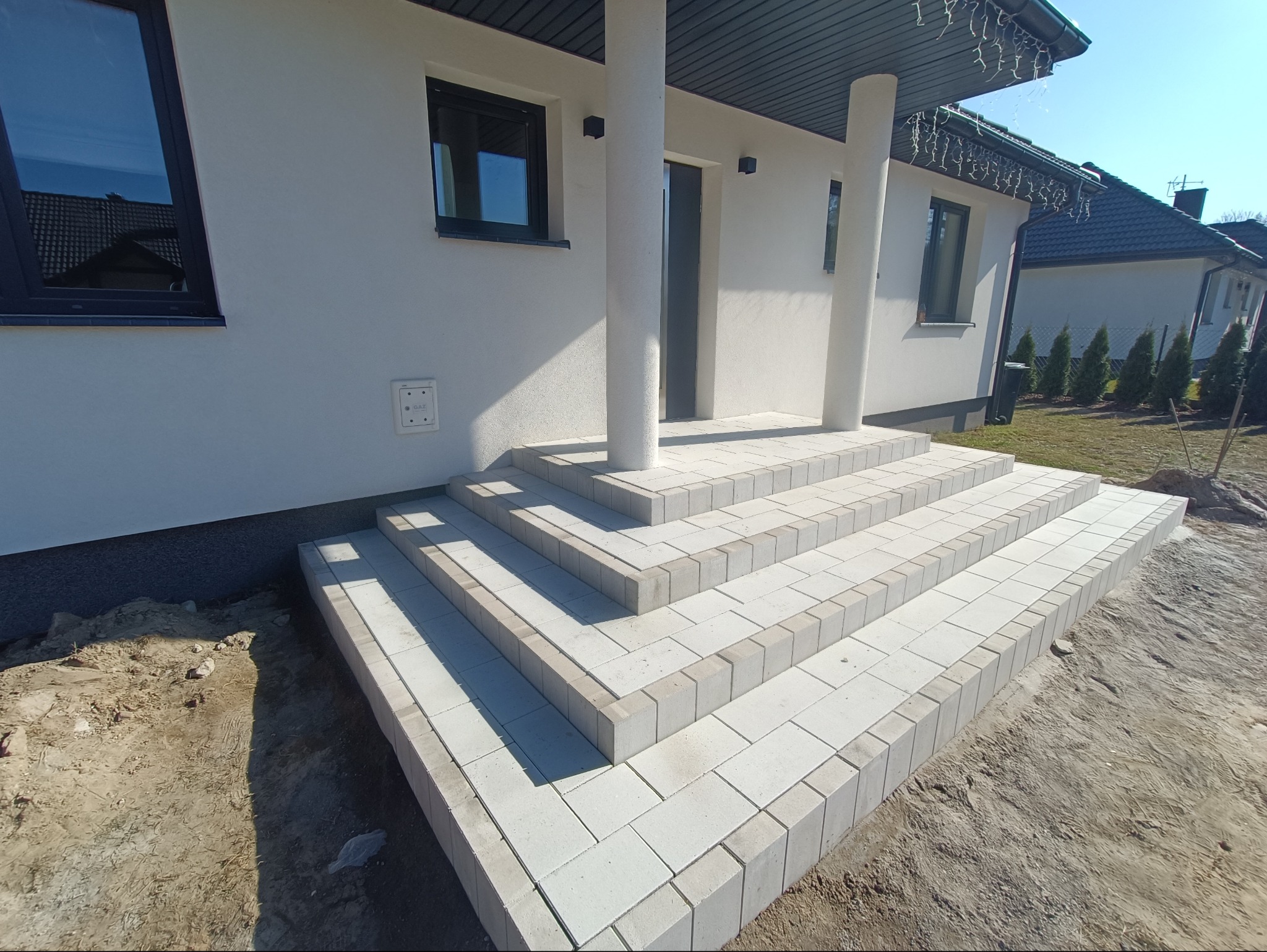 Nowe, jasnoszare schody wejściowe z kostki brukowej prowadzące do nowoczesnego domu z białymi kolumnami.