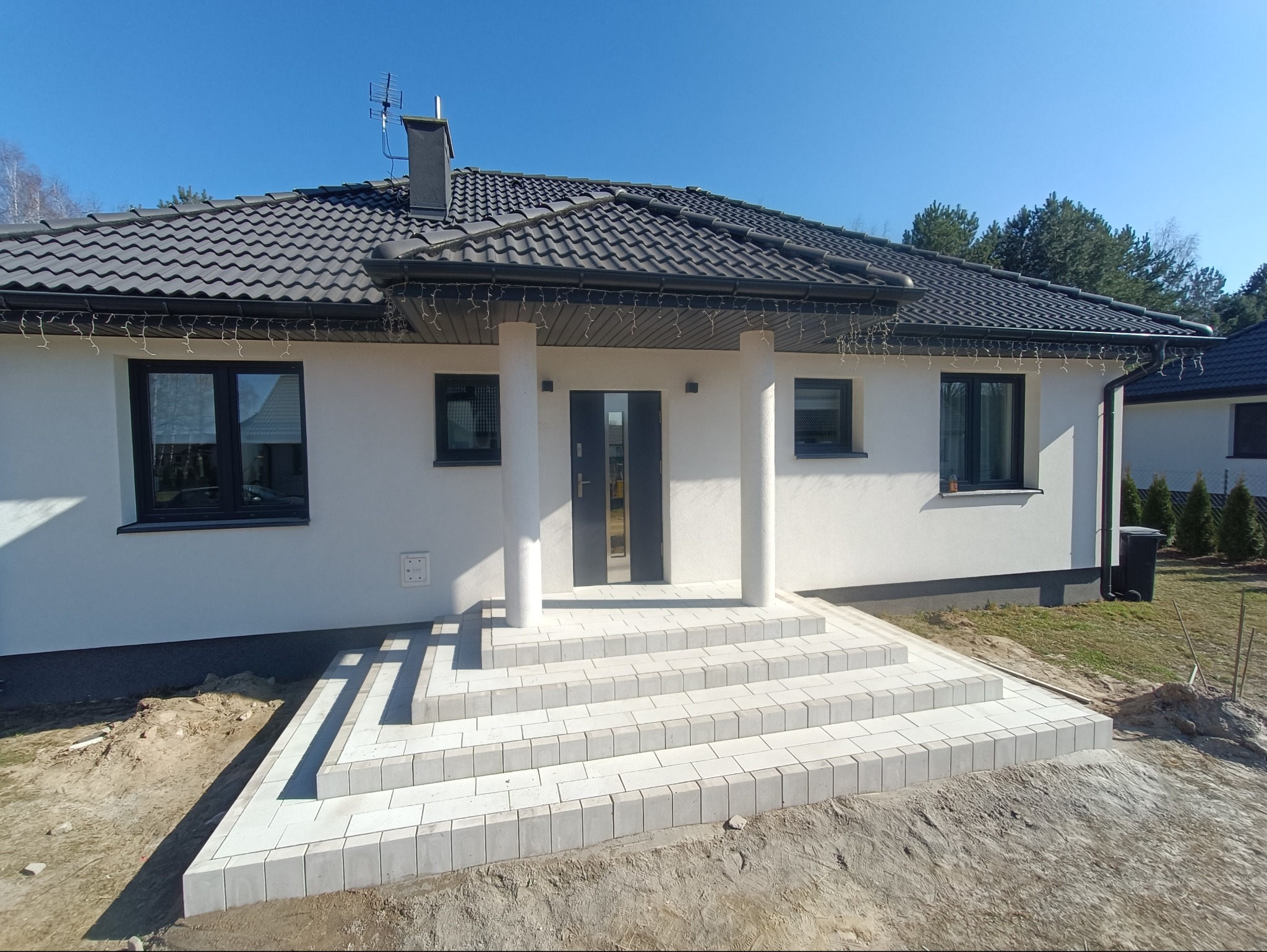 Nowoczesny dom jednorodzinny z wejściem wyłożonym jasnoszarą kostką brukową tworzącą schody, biała elewacja i ciemna dachówka, błękitne niebo w tle.