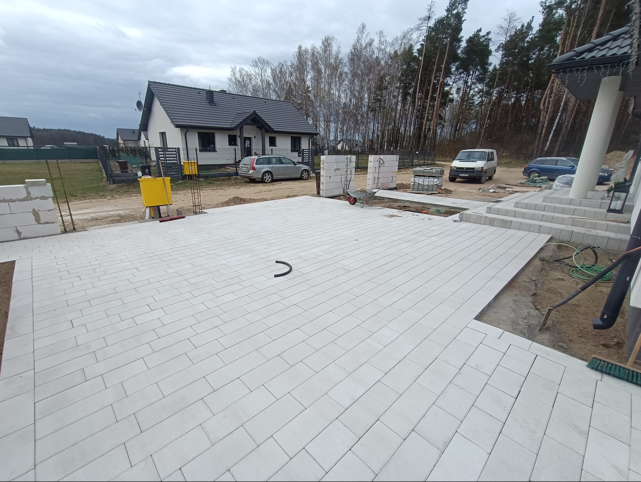 Ułożona jasnoszara kostka brukowa przed domem jednorodzinnym z widocznym fragmentem niezakończonych prac i wystającym elementem rury na powierzchni kostki.