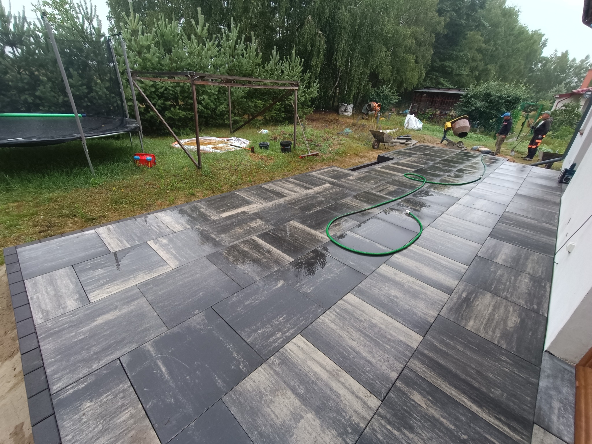Ułożona kostka brukowa w szaro-beżowych odcieniach, wzór jodełki. W tle trawnik, sprzęt budowlany i dwie osoby przy betoniarce. Widok na świeżo wykonany podjazd.