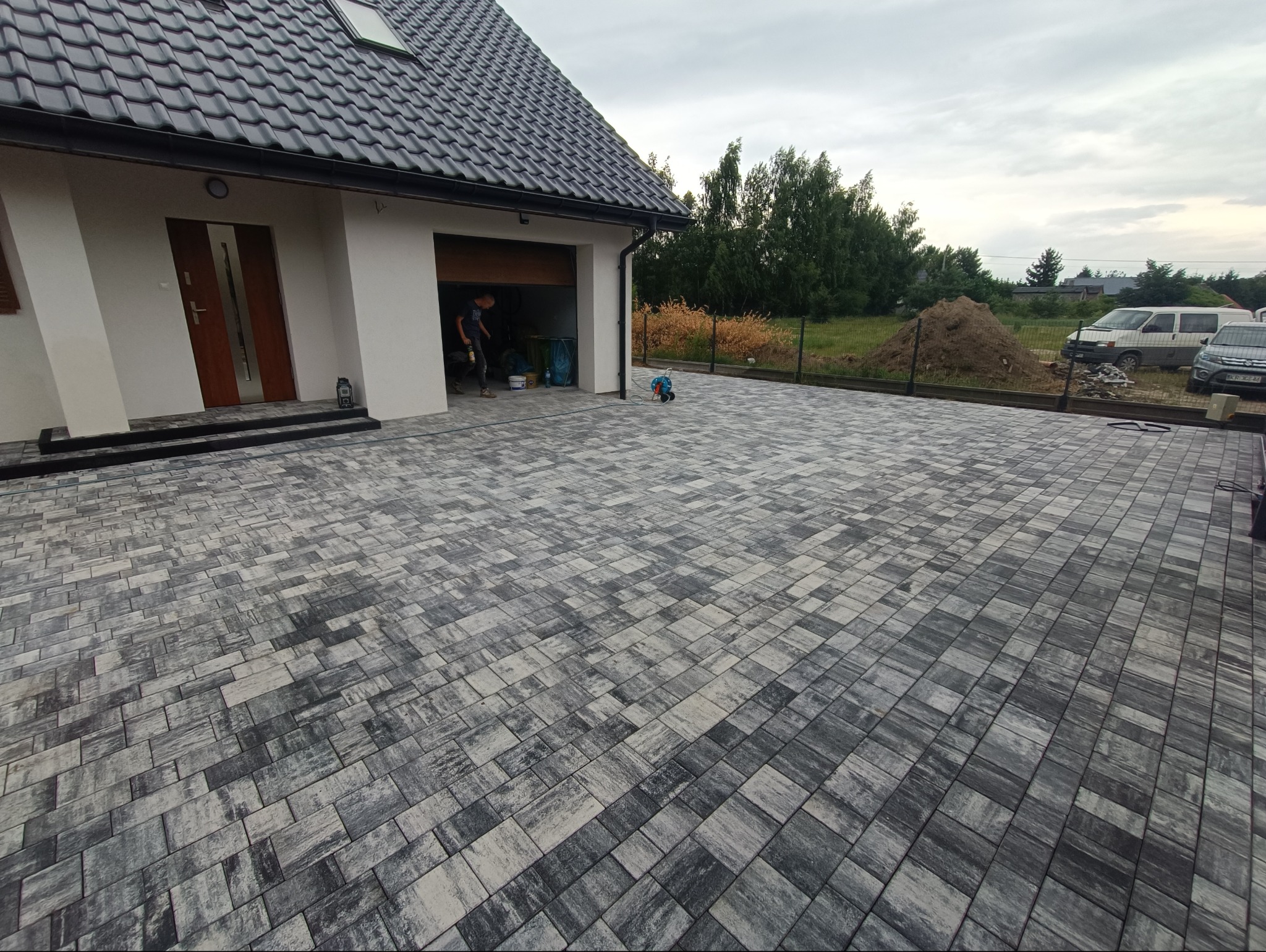 Ułożona kostka brukowa w odcieniach szarości tworzy wzór na podjeździe przed domem z garażem. W tle widoczny ogród i zaparkowane samochody.