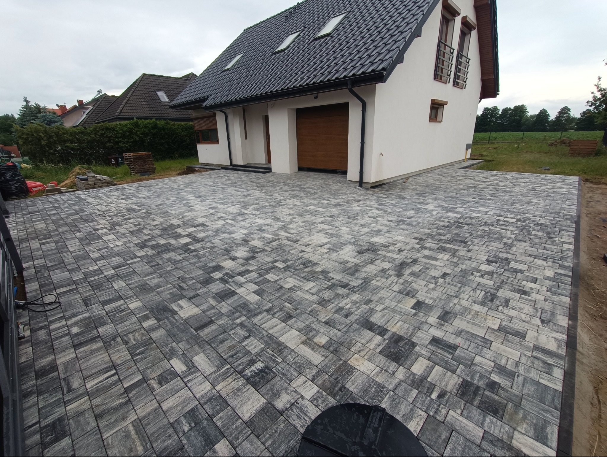 Nowoczesna kostka brukowa w odcieniach szarości tworzy wzór jodełki na podjeździe przed domem z brązową bramą garażową i oknami z ozdobnymi balustradami.