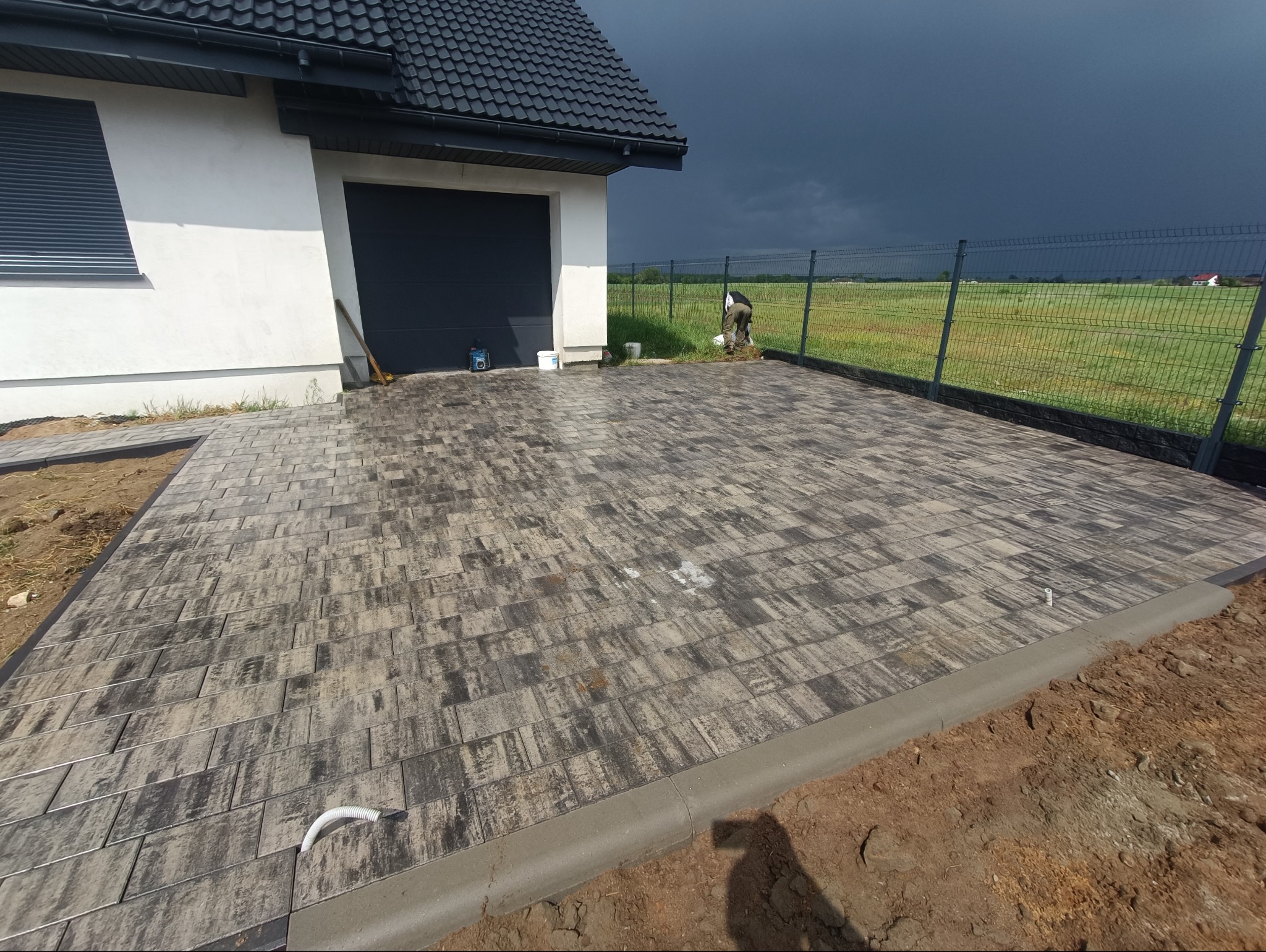 Nowa, szara kostka brukowa ułożona na podjeździe przed domem z garażem, obok metalowe ogrodzenie. Widoczne ciemne chmury na niebie.