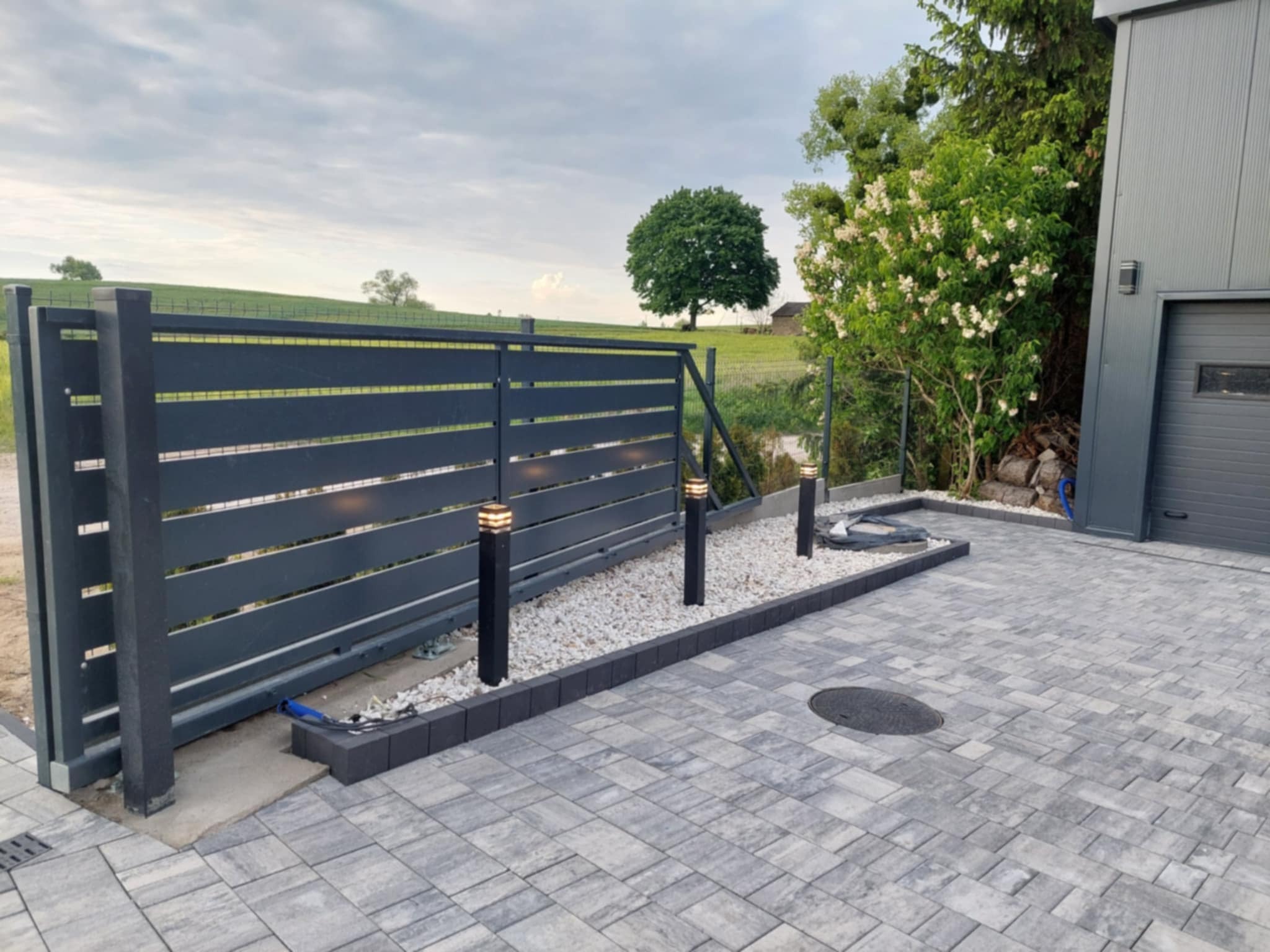 Nowoczesne ogrodzenie panelowe w grafitowym kolorze z subtelnym oświetleniem punktowym, wykończone kostką brukową w odcieniach szarości. Estetyczne połączenie funkcjonalności i designu.