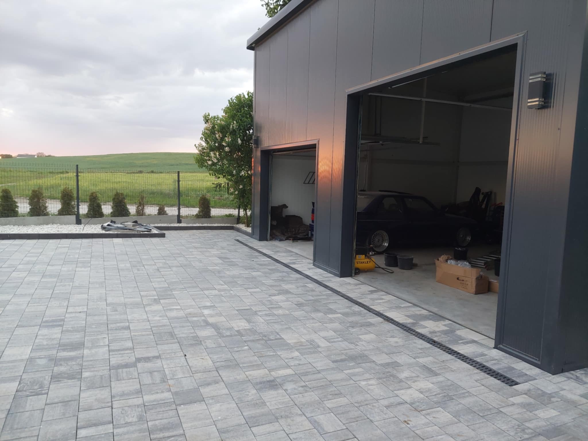 Szara kostka brukowa ułożona przed garażem z metalową elewacją i odpływem liniowym. W tle widoczne zielone pola i drzewa, co dodaje naturalnego charakteru.