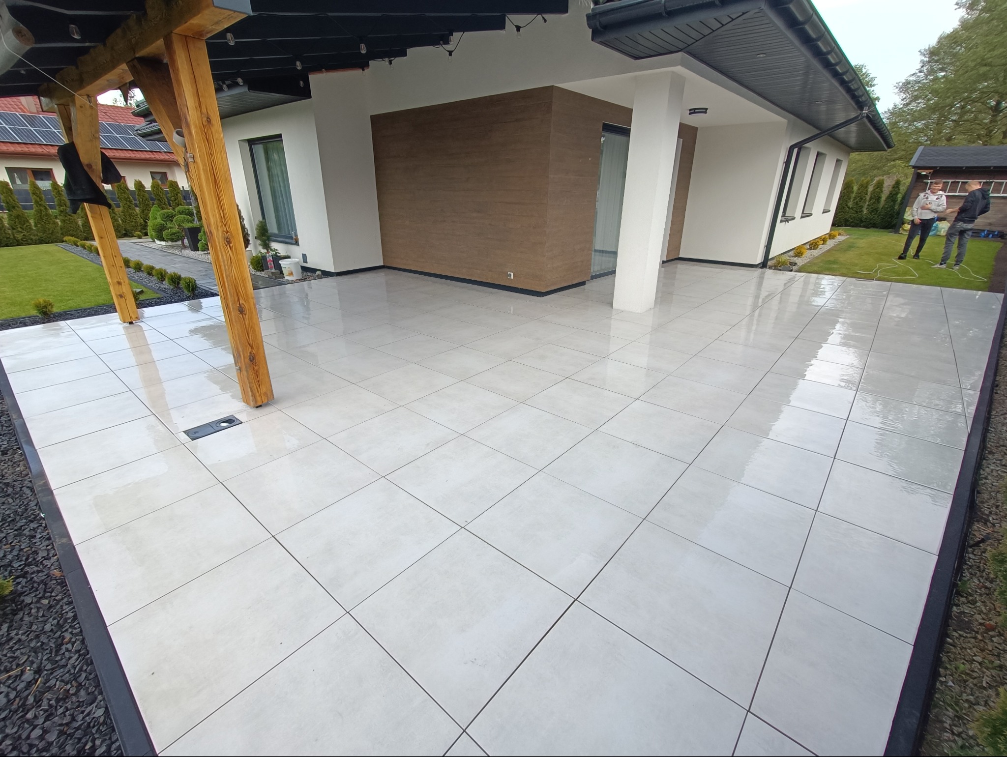 Nowoczesny taras z jasnych, kwadratowych płytek, drewniana pergola, czarne obrzeże, widok na dom i ogród. Dwóch mężczyzn w tle.