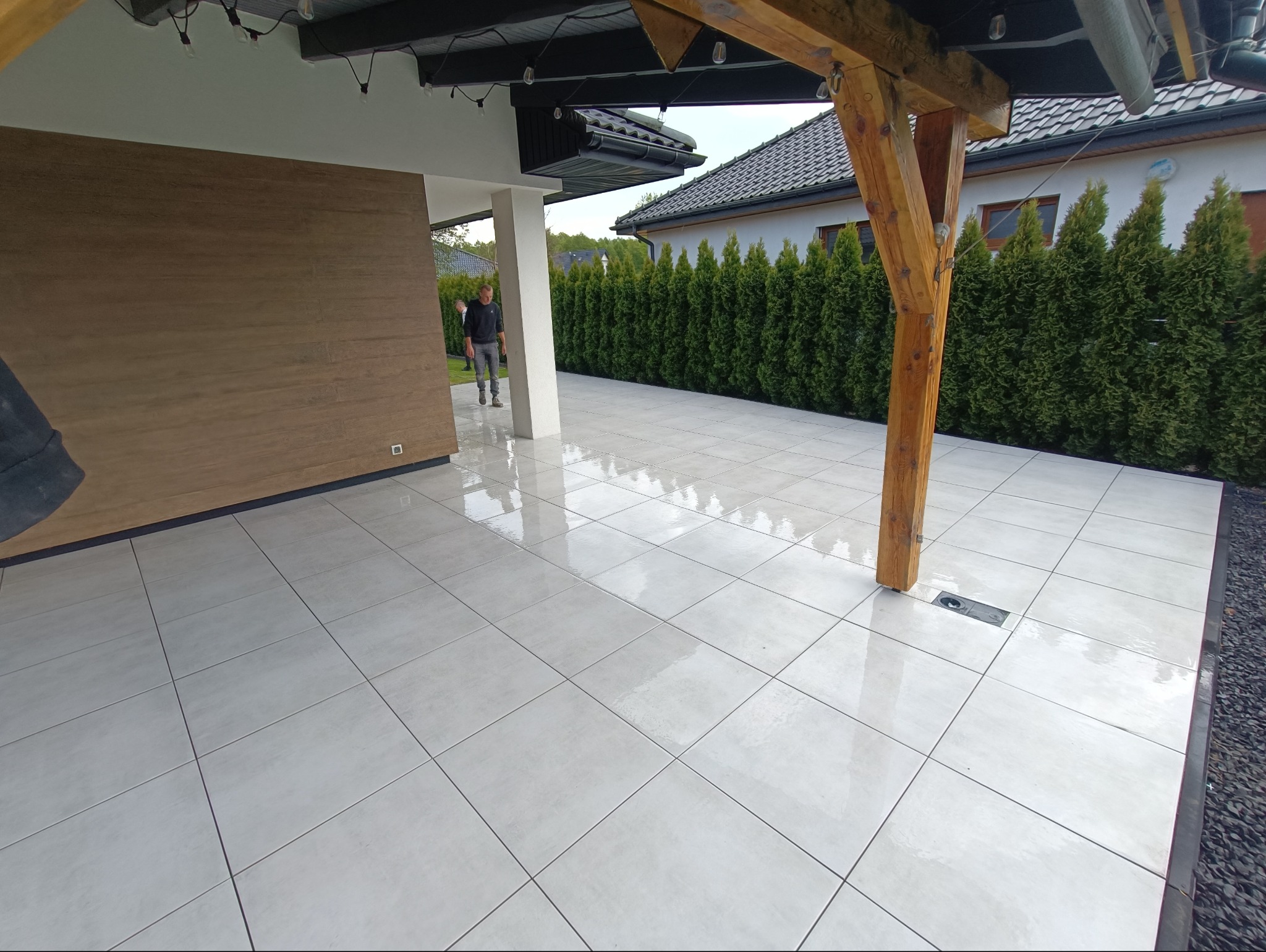 Białe płytki tarasowe ułożone na zewnątrz budynku z drewnianą konstrukcją i zielonymi tujami w tle. Widoczny odpływ liniowy.