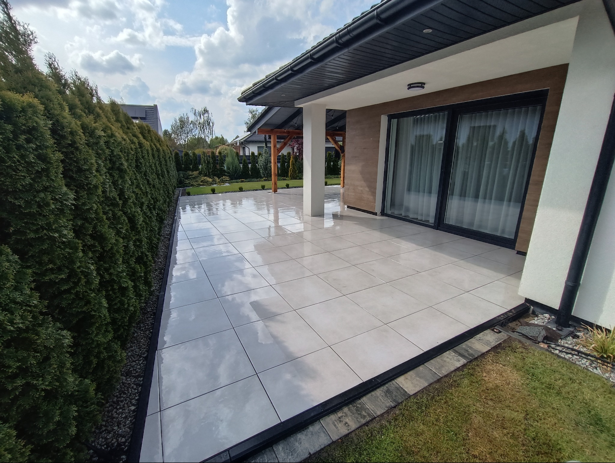 Jasnoszara, błyszcząca, kwadratowa podłoga z płytek na zewnątrz domu. Widoczny fragment ogrodu i żywopłot. Nowoczesny design i dbałość o detale.