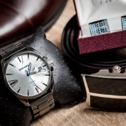 Zestaw męskich akcesoriów: srebrny zegarek Diesel na czarnej poduszce, skórzany pasek Pierre Cardin z metalową klamrą, spinki do mankietów w pudełku oraz brązowe skórzane buty w tle.