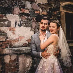 Para młoda pozuje na tle starej, ceglanej ściany z widocznymi śladami renowacji; panna młoda w koronkowej sukni z welonem, pan młody w szarej marynarce w kratę.