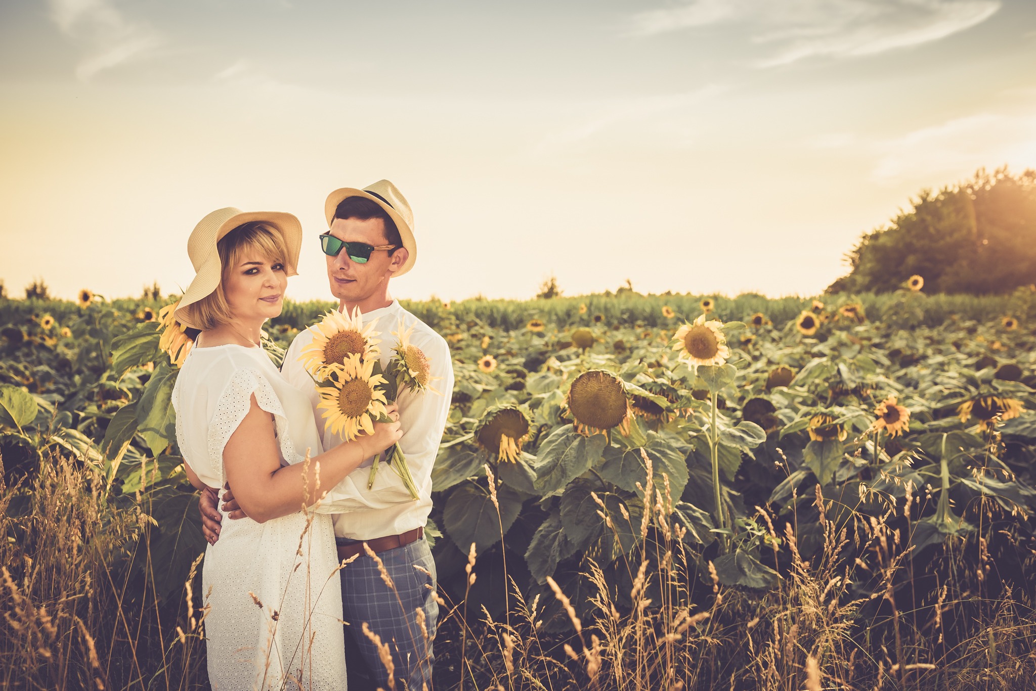 Para zakochanych w kapeluszach i okularach przeciwsłonecznych, obejmująca się na polu słoneczników, trzyma bukiet słoneczników.