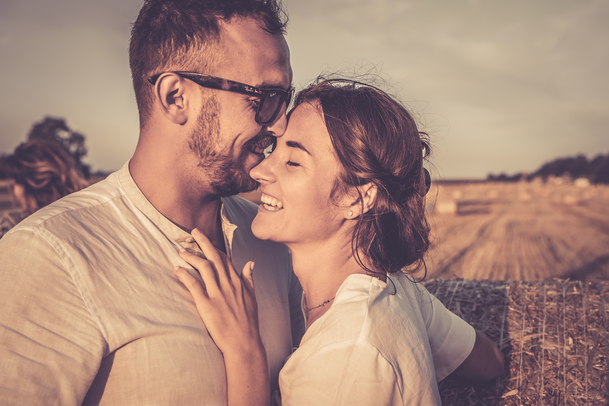 Uśmiechnięta para w objęciach na tle pola ze słomianymi balotami, mężczyzna w okularach Ray-Ban, ciepłe światło.