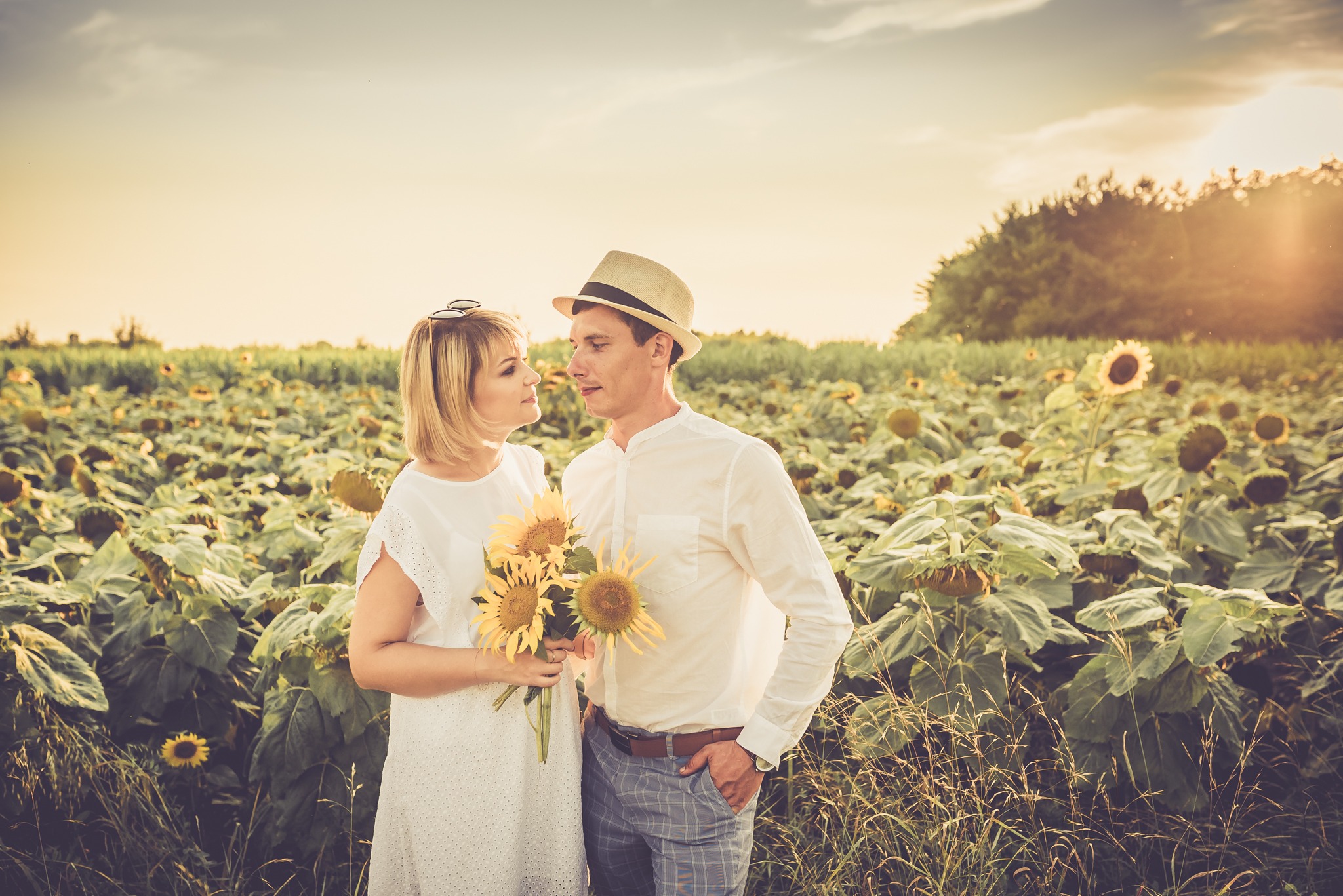 Para wpatrująca się w siebie na polu słoneczników podczas zachodu słońca, kobieta trzyma bukiet słoneczników, oboje ubrani w jasne stroje.